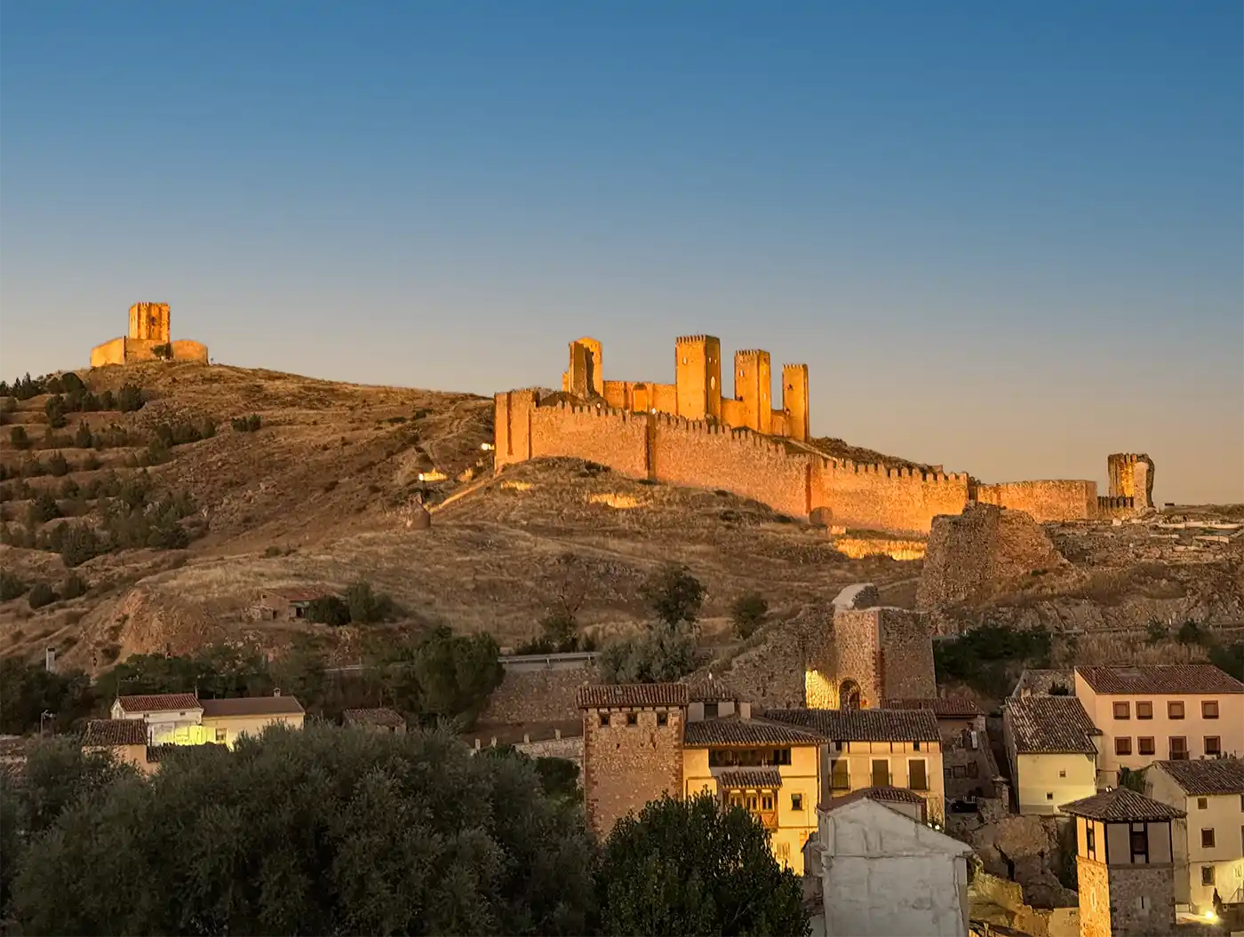 Molina-de-Aragon-Burg-beleuchtet Eine alte Burganlage aus Stein auf einem Hügel oberhalb einer kleinen Stadt bei Sonnenuntergang.