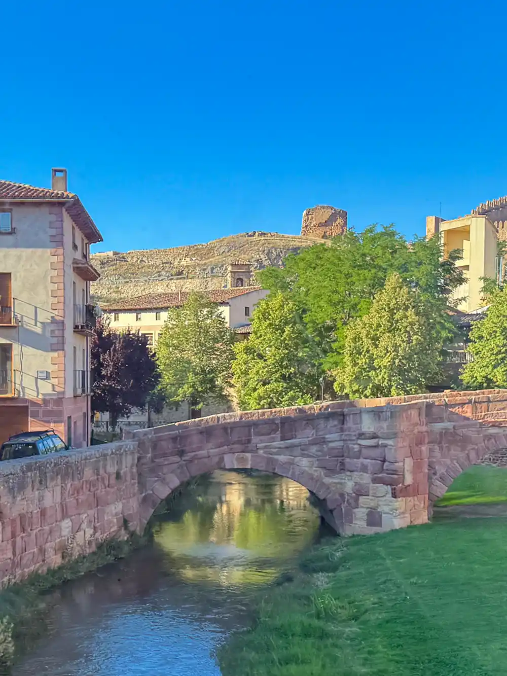 Molina-de-Aragon-Puente-Viejo-links Eine Steinbrücke über einen kleinen Fluss, mit Häusern, Bäumen und einer Ruine auf einem Hügel unter strahlend blauem Himmel.
