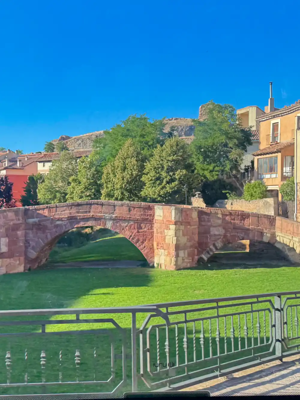 Molina-de-Aragon-Puente-Viejo-rechts Eine Steinbrücke über grünem Gras, Bäumen und alten Gebäuden unter einem strahlend blauen Himmel.