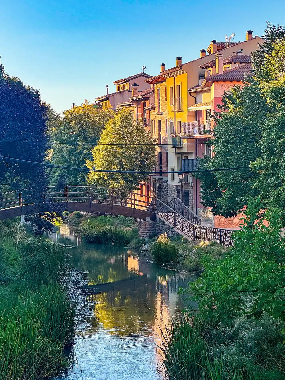 Molina-de-Aragon-Rio-Gallo Eine hölzerne Fußgängerbrücke, die vor farbenfrohen Gebäuden über einen Bach führt.