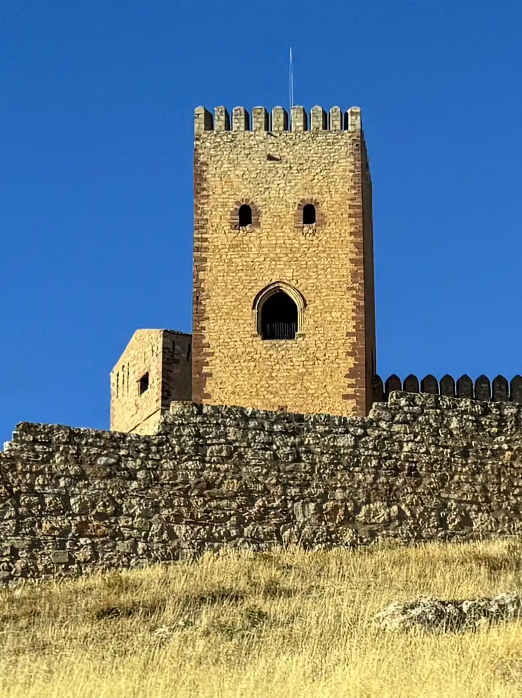 Molina-de-Aragon-Torre-Dona-Blanca Der Steinturm einer Festung ragt über einer Umfassungsmauer vor einem strahlend blauen Himmel empor.