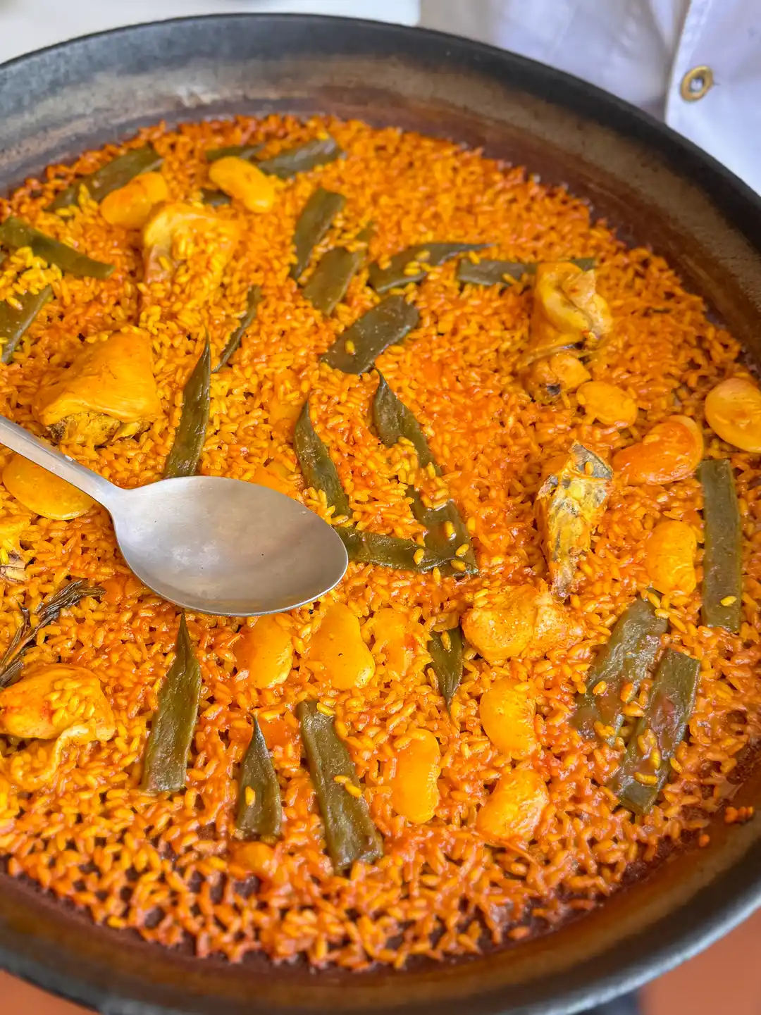 Eine große Pfanne mit Hähnchen-Bohnen-Paella, auf der ein Metalllöffel liegt.