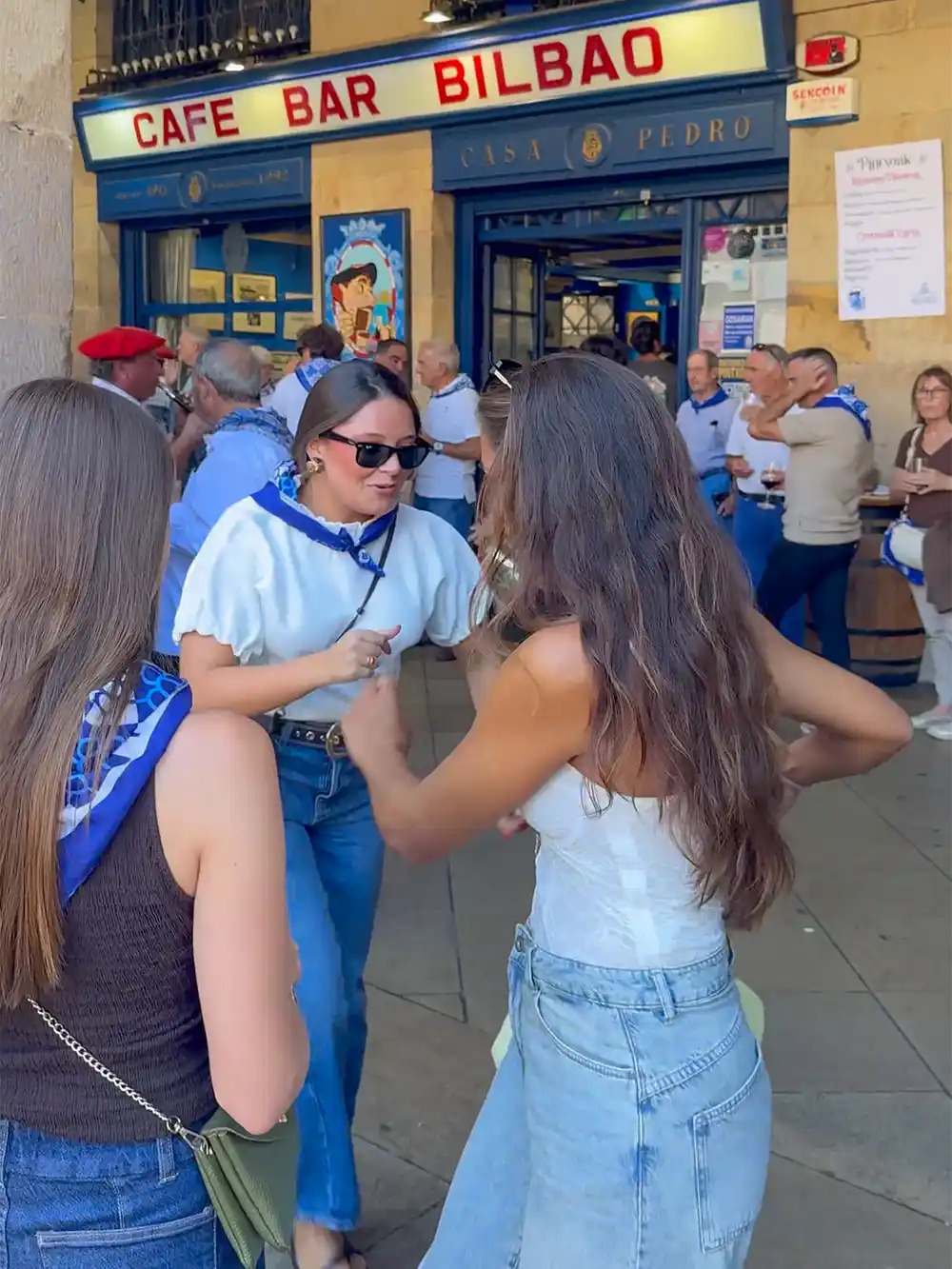 Drei Frauen unterhalten sich und lachen vor einem belebten Café, vor dessen Eingang sich Menschen versammelt haben.