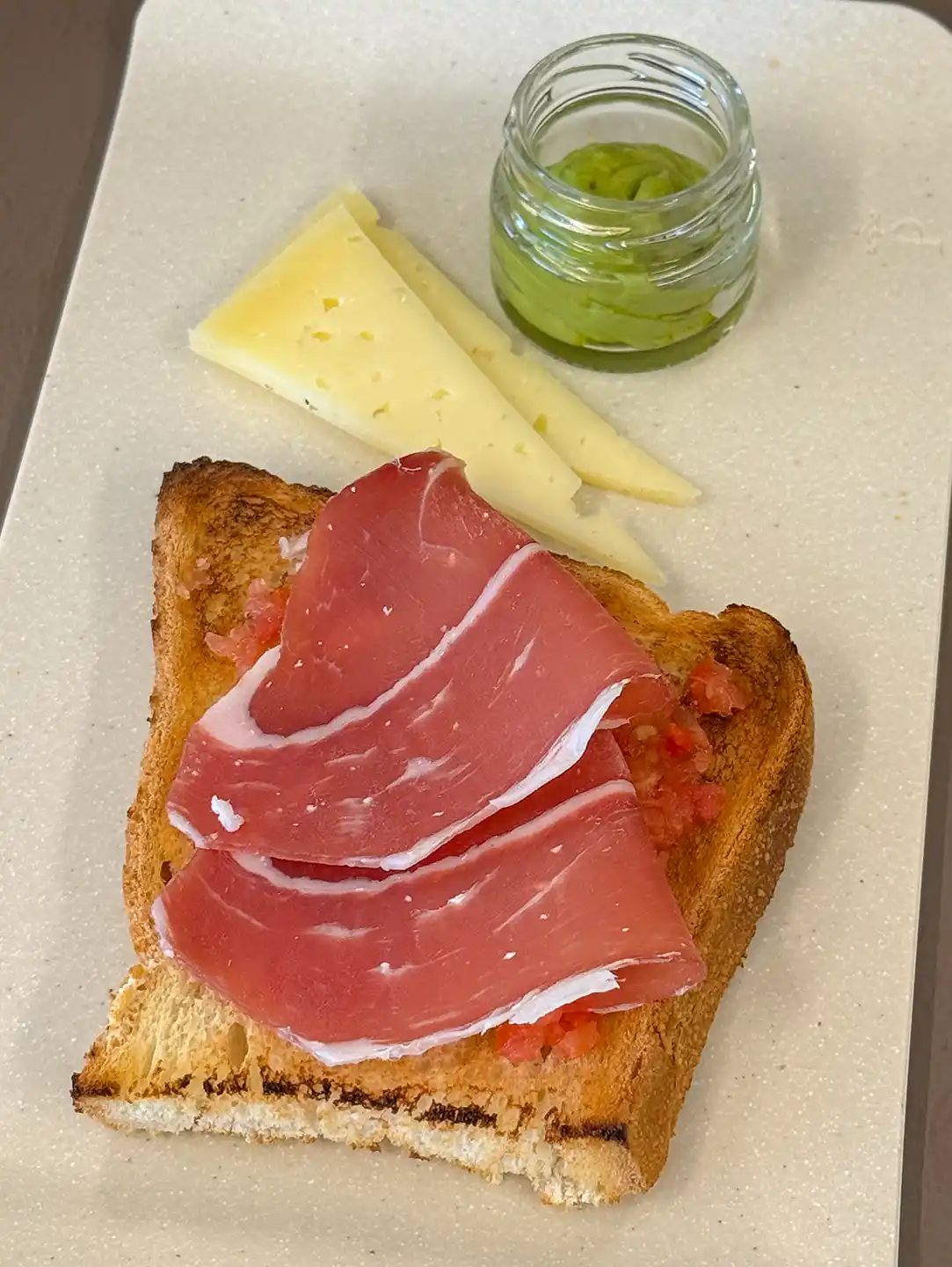 Toast mit Tomate und spanischem Schinken, serviert mit zwei Käsescheiben und einem kleinen Glas grüner Avocadocreme.