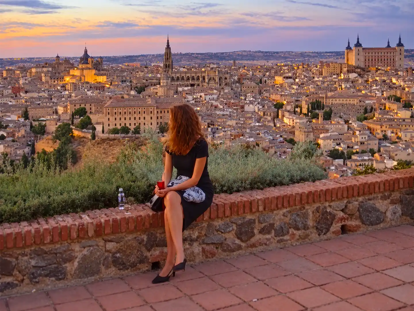 Toledo-Panorama-Mirador-del-Valle Eine Frau sitzt bei Sonnenuntergang auf einer Steinmauer mit Blick auf die Stadt Toledo.