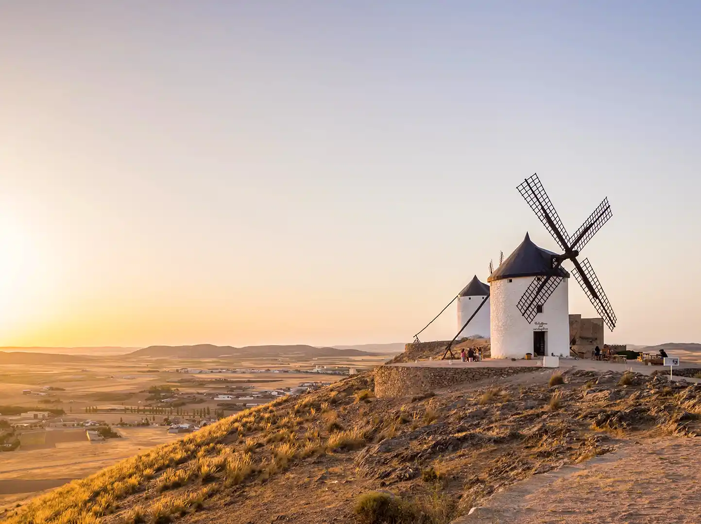 Windmuehlen-Kastilien-La-Mancha Zwei Windmühlen auf einem Hügel mit Blick auf ein Tal bei Sonnenuntergang.
